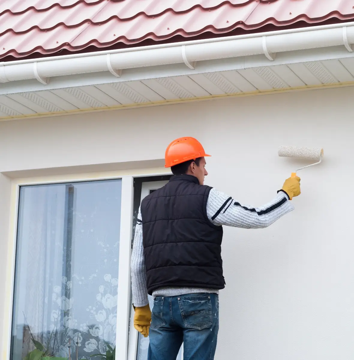 Person in a hard hat and gloves painting a house exterior wall with a roller under a tiled roof.