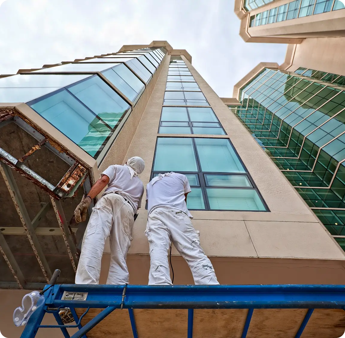 Two workers in white uniforms stand on scaffolding, looking up at a tall building with large glass windows, under a cloudy sky.